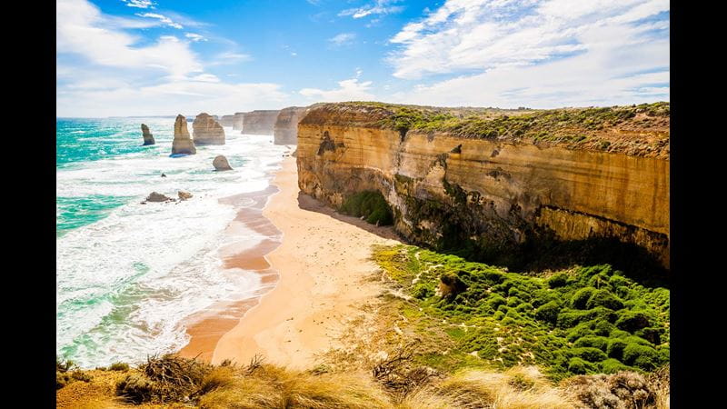 A view over cliffs above a sandy beach with rock formations out to see and waves rolling in, in Australia, with the sun shining