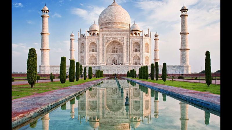 The ivory white marble front of the Taj Mahal building, in India, with its water feature in the foreground and blue sky behind
