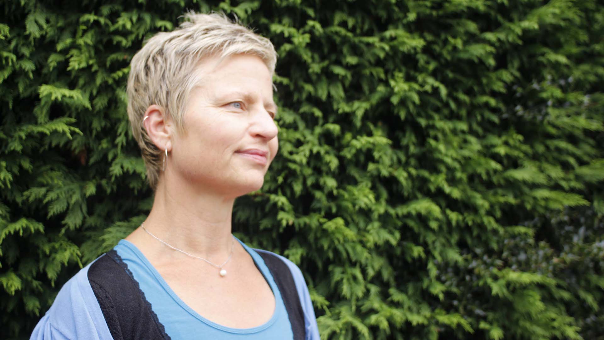 Headshot of Suzi Griffin, yoga instructor; a woman in a blue t-shirt with cropped blonde hair against a leafy background