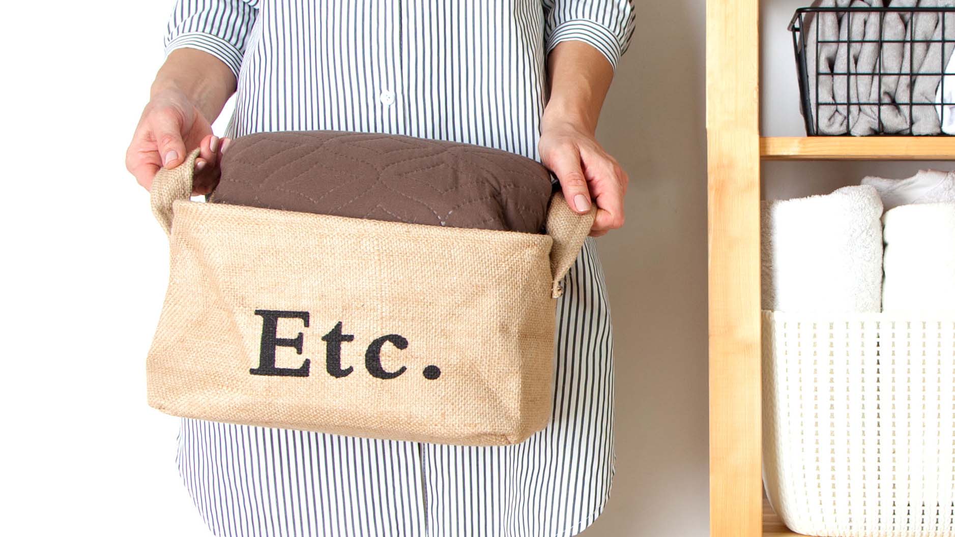 A person wearing a striped shirt holding a basket which has 'Etc.' printed in black letters and contains a brown cushion.
