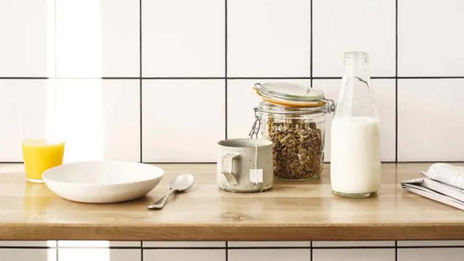 A wooden counter topped with a white bowl and glass of orange juice, along with a jar of cereal, jug of milk and a mug. The backdrop is white tile with grey grouting.