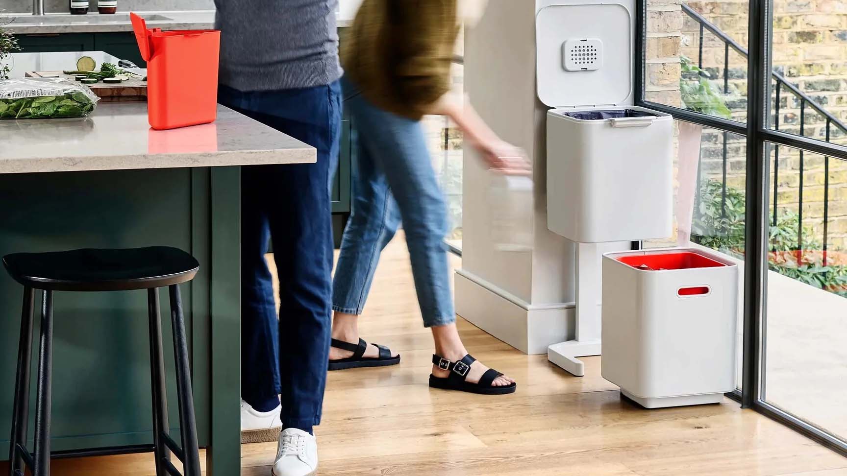 Two people preparing food a kitchen; one is standing at a large island while the other person places an item in a pedal bin.