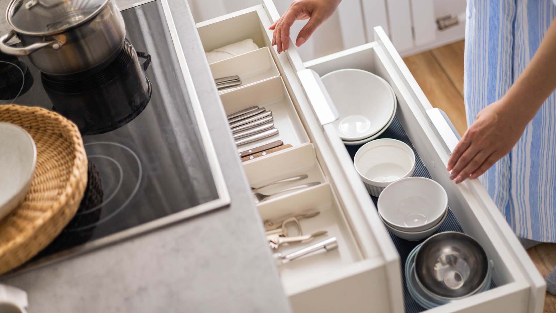 A pair of hands opens two kitchen drawers containing neatly organised cutlery and a selection of bowls and other kitchen items.