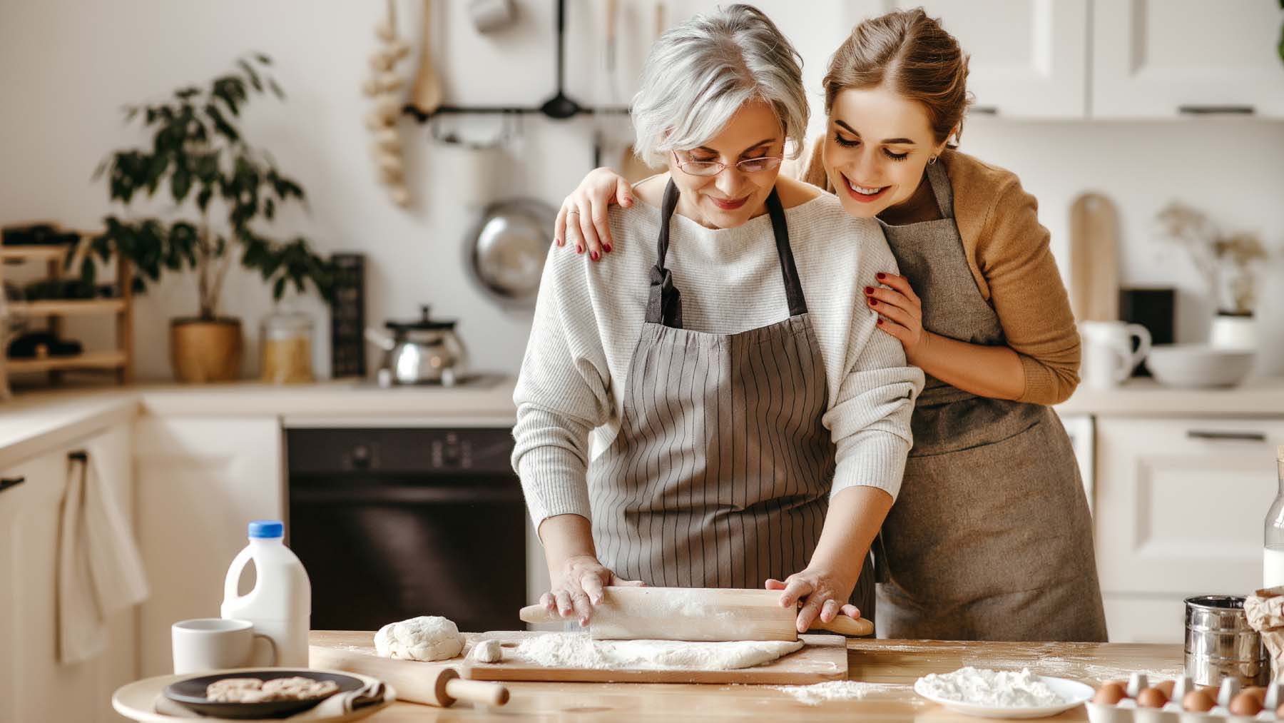 Two woman baking together; a silver-haired woman uses a rolling pin at a kitchen counter while a younger woman affectionately holds her shoulders.