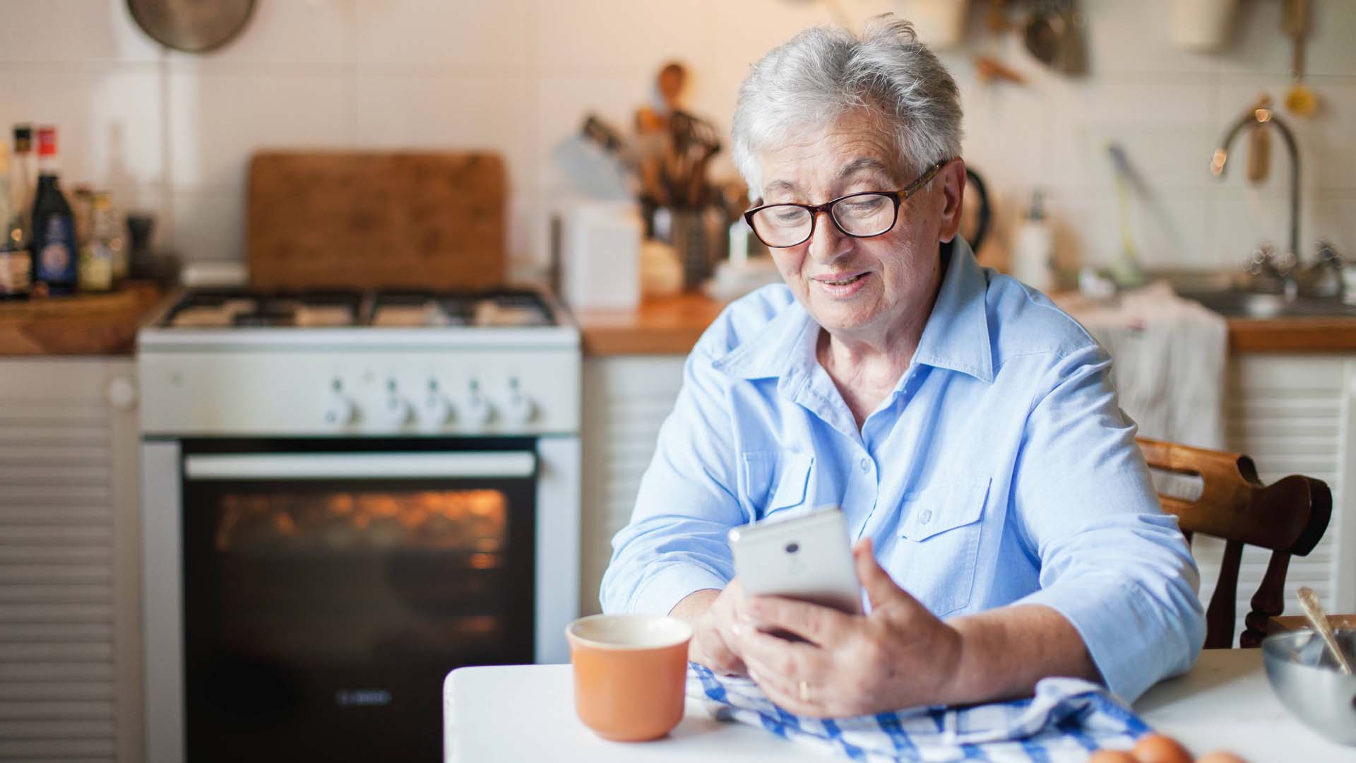 An older person sitting at a kitchen table using a mobile phone.