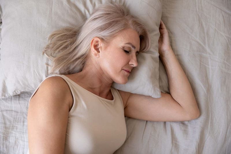 Head and shoulders of a middle-aged woman asleep in bed with her head on a pillow.