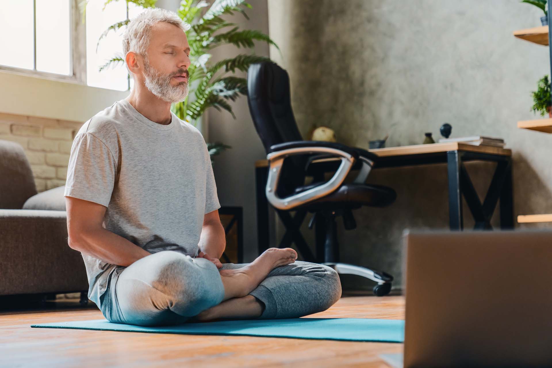 A middle-aged man with short grey hair and facial hair sits cross-legged on a blue yoga mat with hands in his lap and eyes closed; a desk, office chair, couch and house plant are visible behind. 