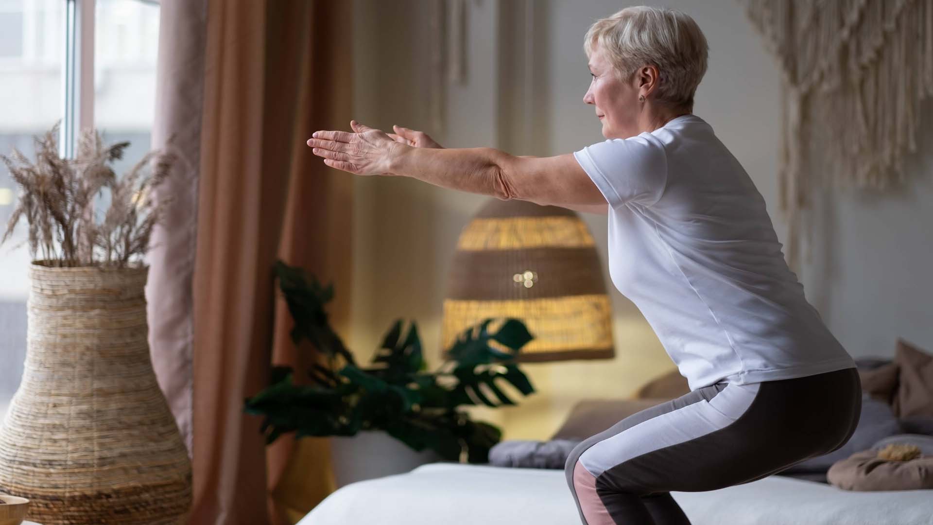 A woman in sports leggings and a white t-shirt practises the chair position in yoga in the comfort of her home