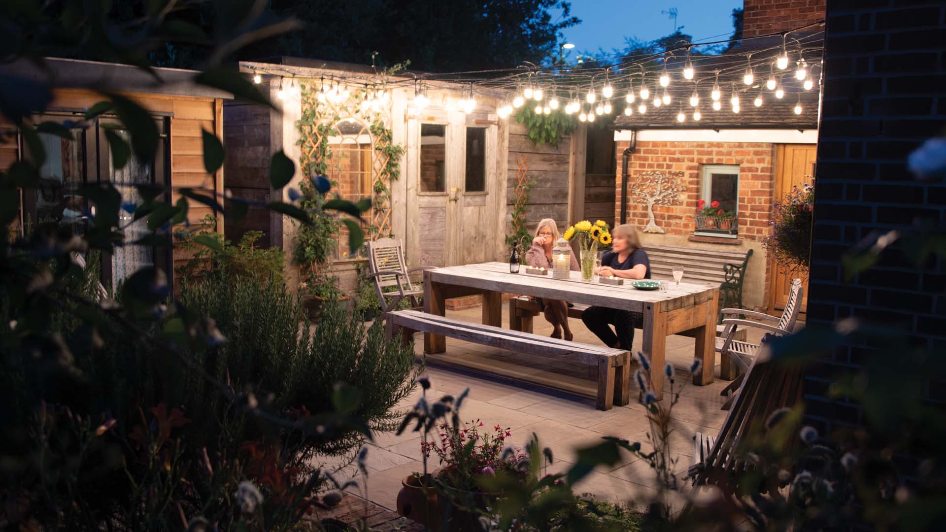 Two women sat in a courtyard garden at night at a large tab;e with bench seating