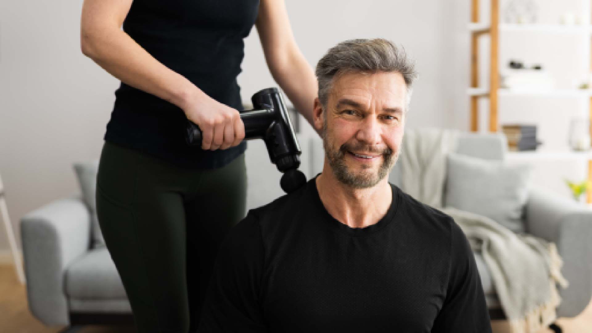A man with grey facial hair enjoys a massage gun being used on his shoulders
