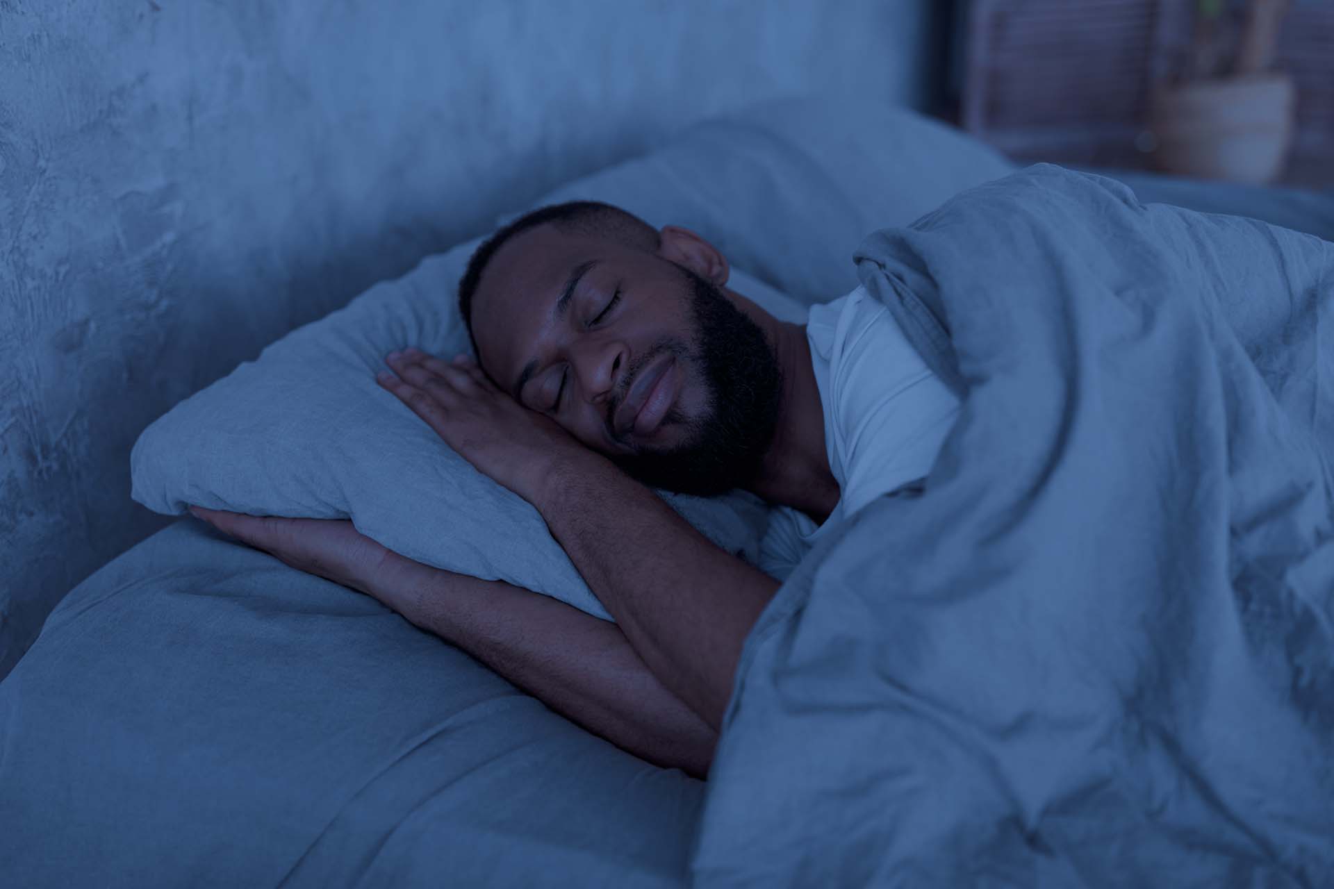 A bearded man sleeping peacefully in bed with his hands on the pillow and a duvet over his shoulders