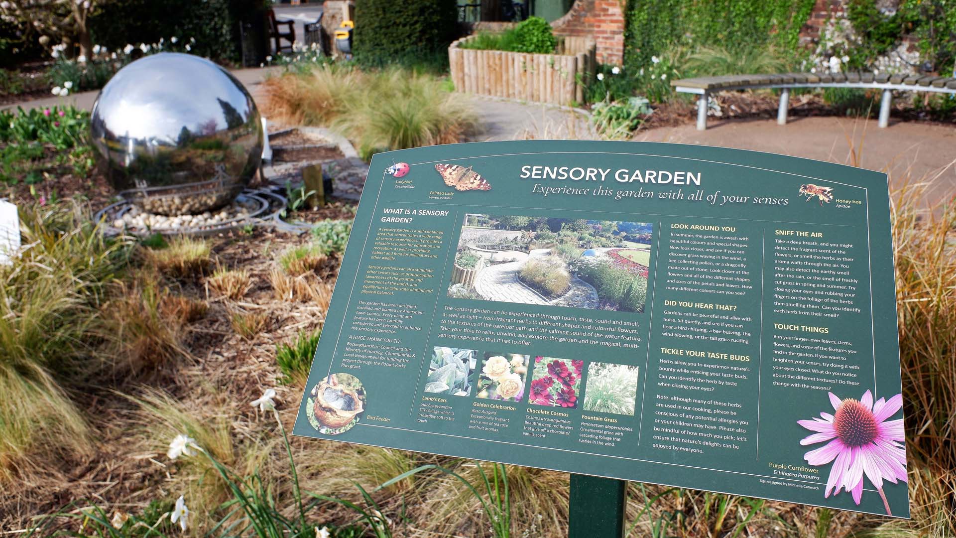 An interpretation board in a sensory garden with planted borders and installations such as a large silver ball.