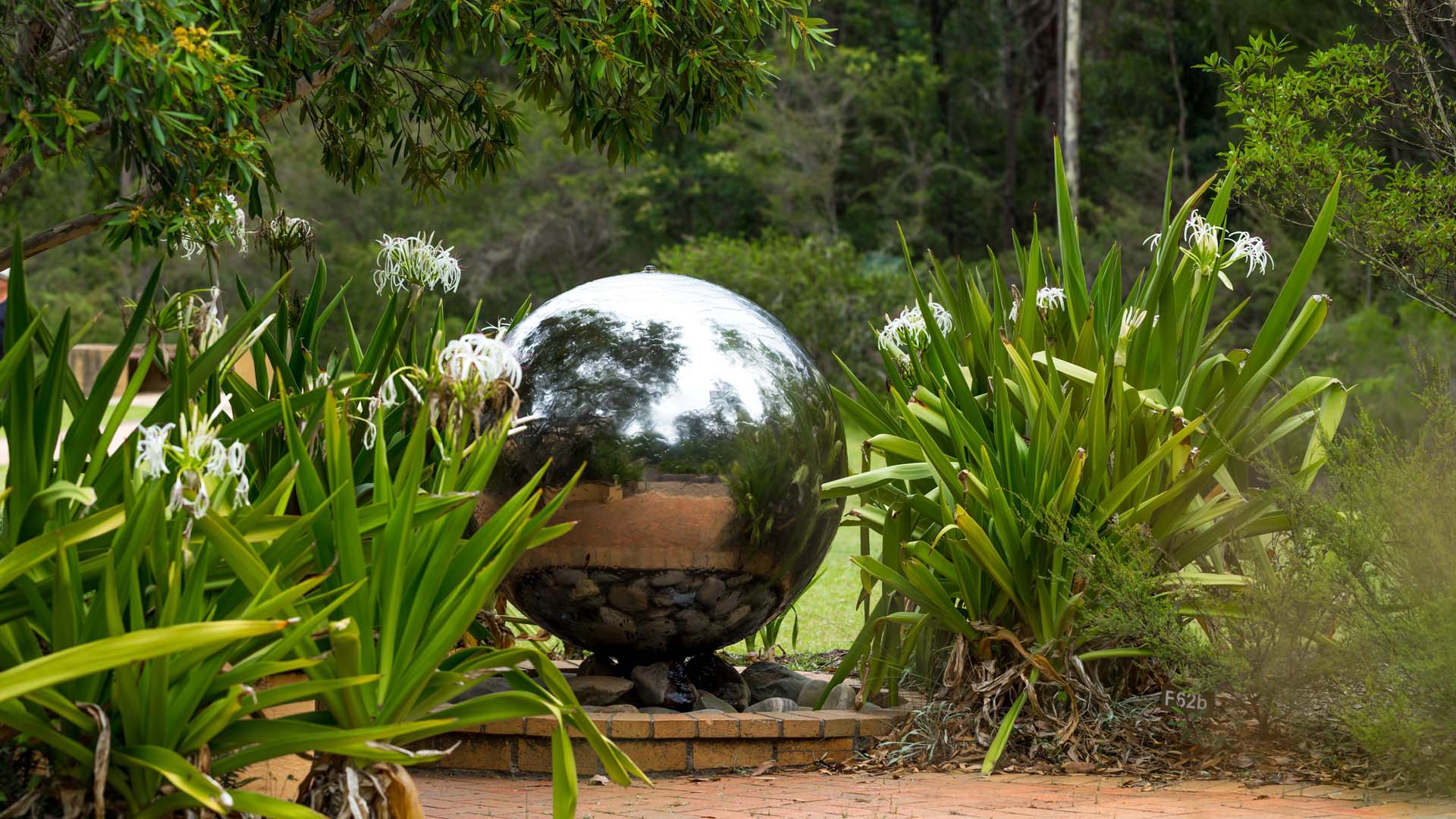 A large silver sphere as part of a water feature in a garden with greenery on both sides.