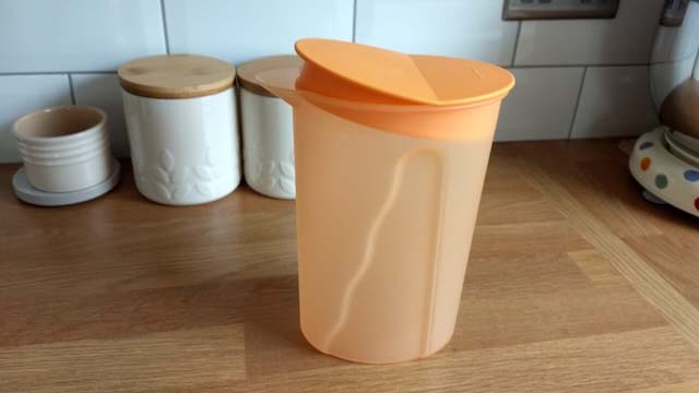 A clear plastic Tupperware jug with an orange lid on top of a wooden kitchen counter, with a row of white kitchen containers against a tiled wall.