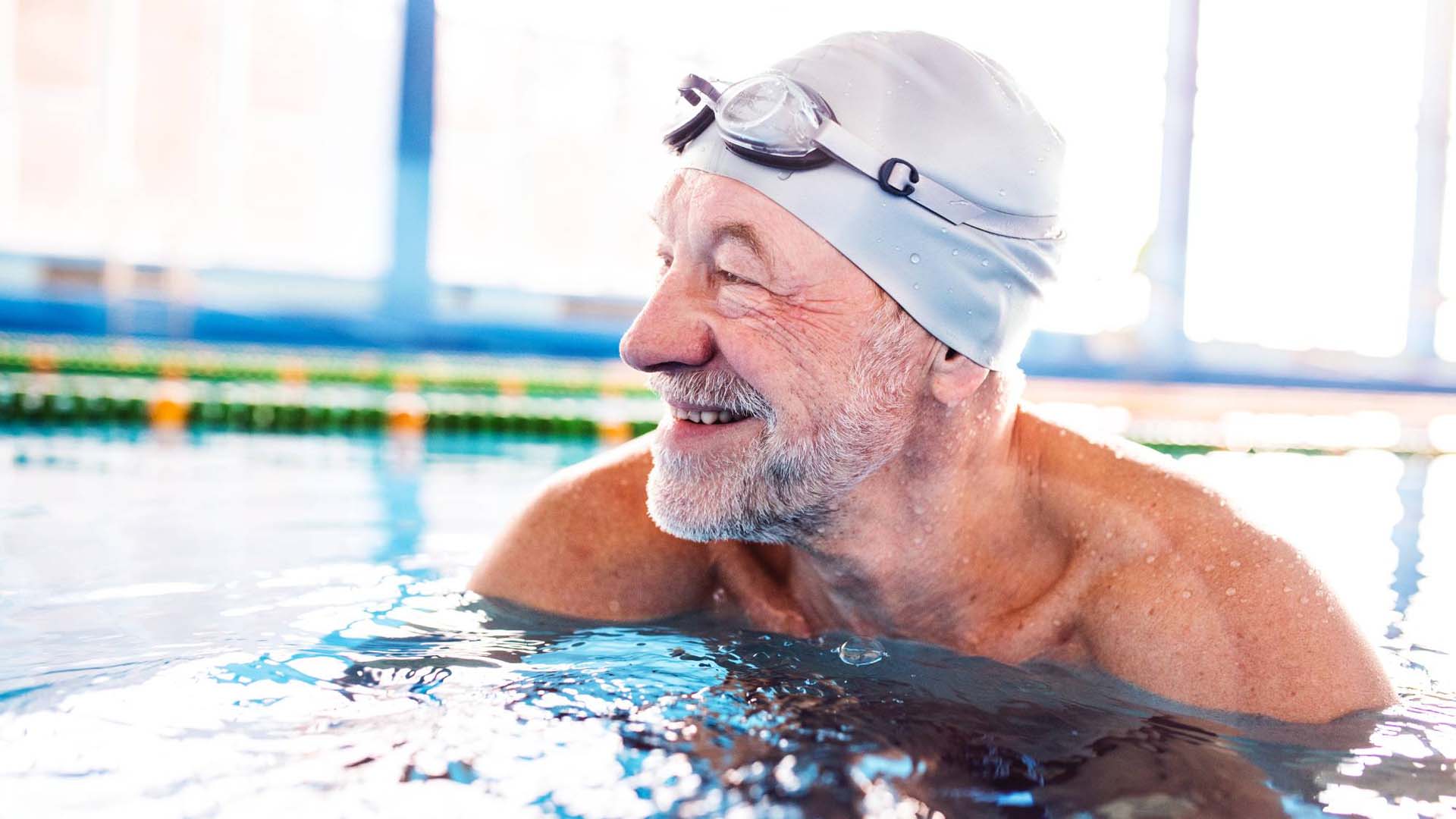 An older man with facial hair wearing a swimming cap and goggles in an indoor swimming pool