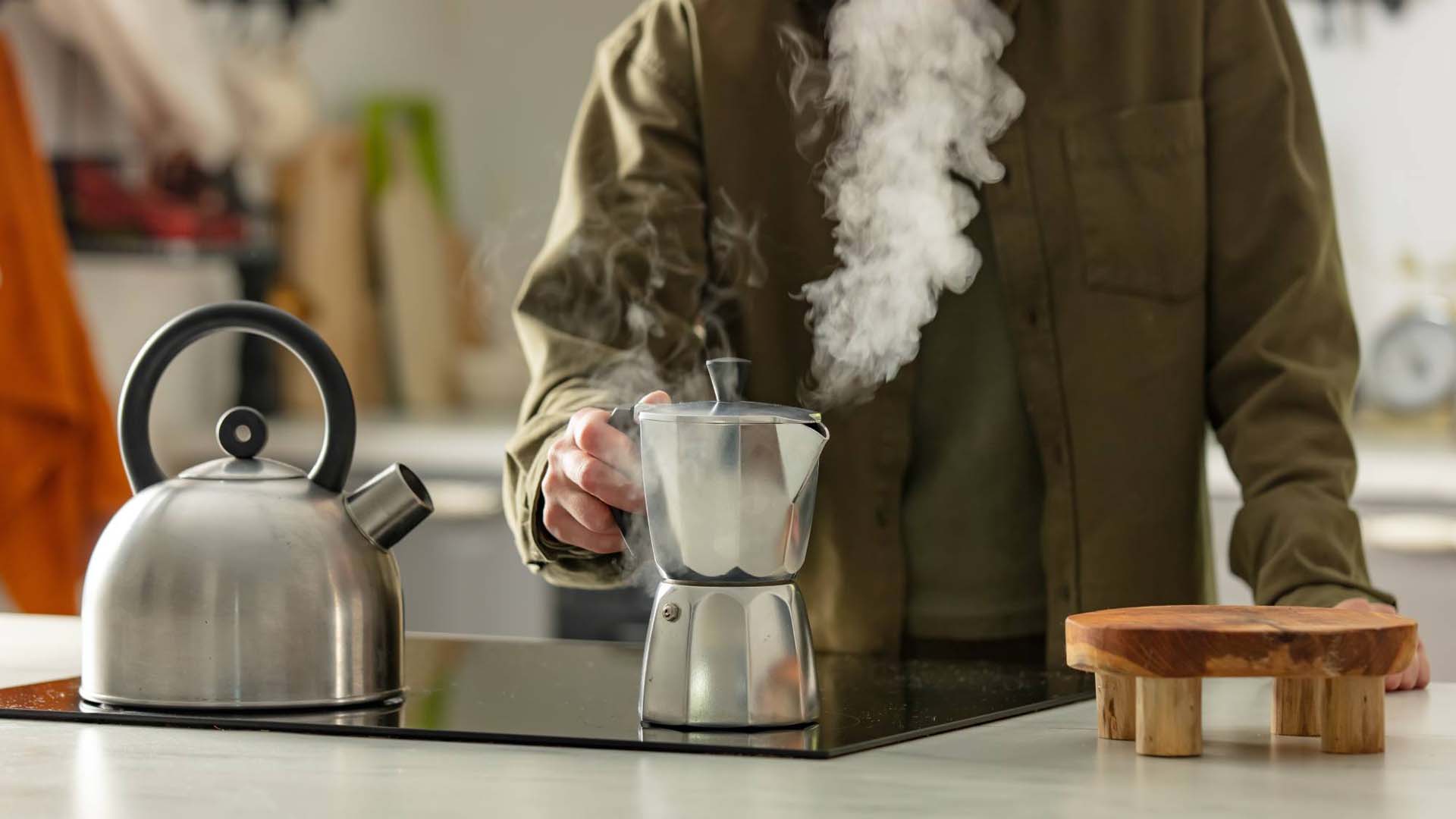 A man standing at a flat hob holding the handle of a steaming silver Moka pot next to a silver kettle.