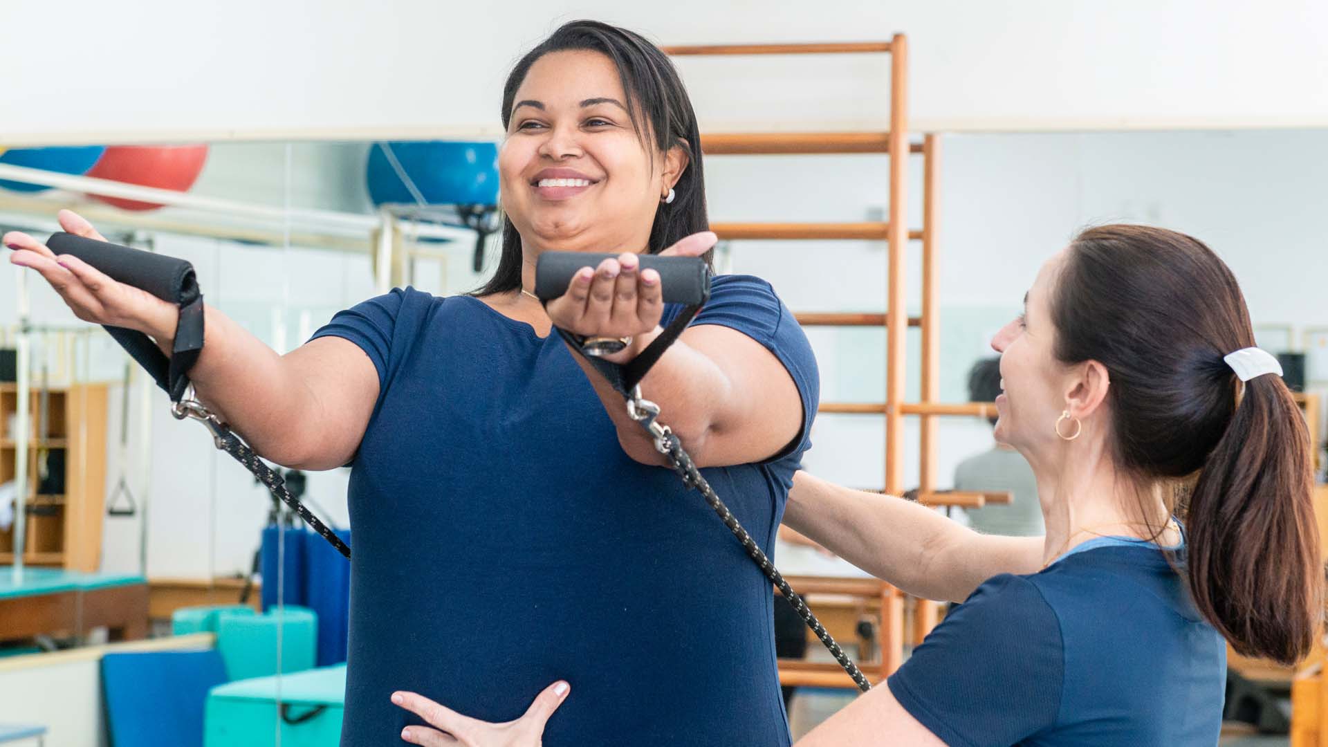 A smiling woman using a reformer Pilates machine with the help of an instructor.