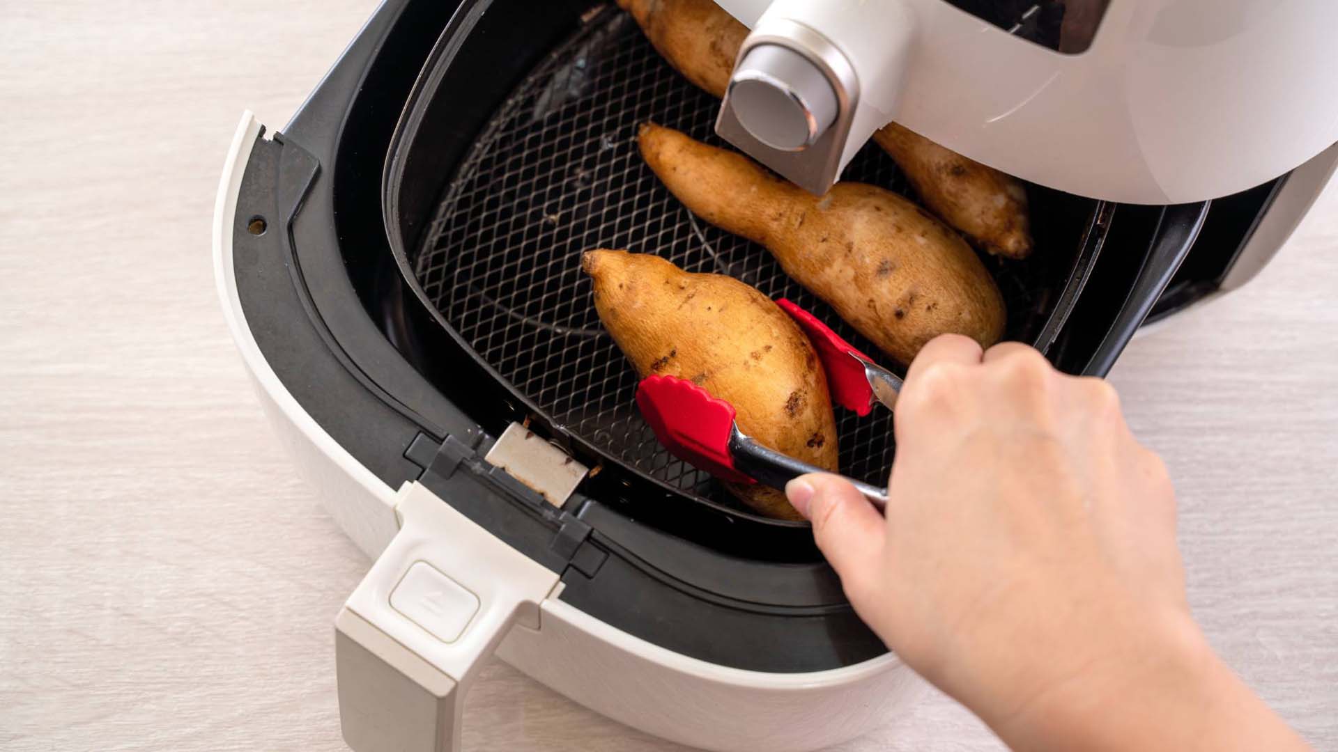 An open air fryer drawer containing potatoes and a pair of red tongs lifting one out