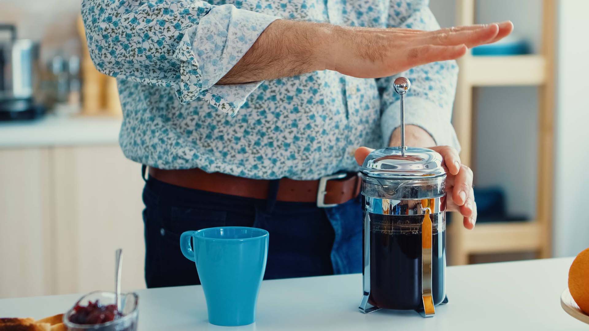 A man wearing a patterned blue shirt depresses a cafetiere of black coffee which stands on a counter next to a blue mug.