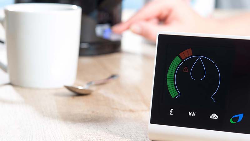 A smart meter is displayed on a  wooden surface near mug and spoon and a kettle which is being switched on by a hand. The meter is giving a digital reading of energy consumption.