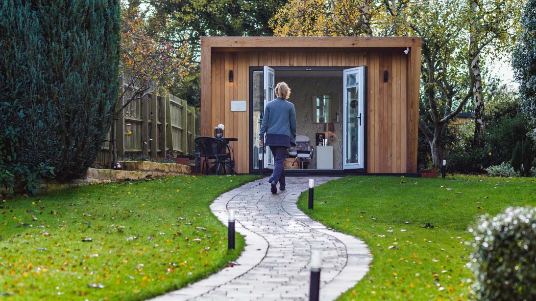 A garden lawn with a brick path, where a woman walks towards a modern garden room.