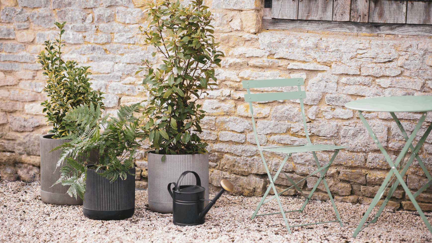 Chic bistro-style chairs on a gravel path with 3 plants pots in front of a old brick light wall