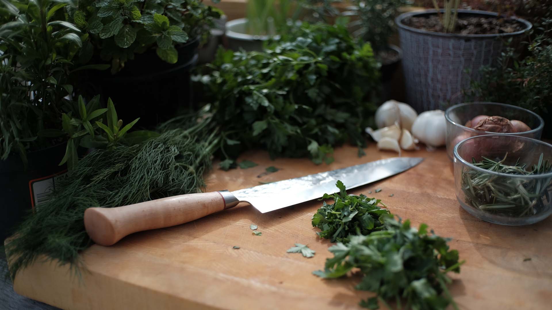 A sharp knife on top of a wooden board surrounded by various fresh herbs