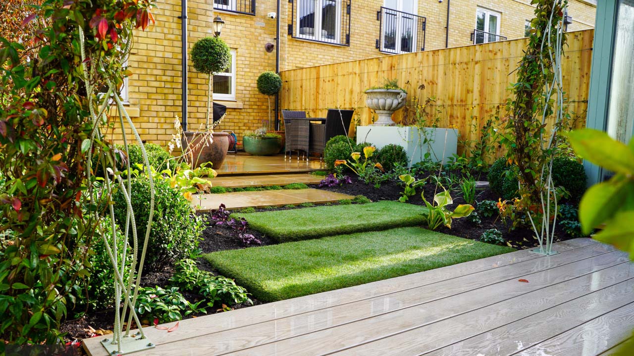 A sloping garden made up of small lawned areas and a decked terrace, surrounded by planted borders with a house visible in the background.