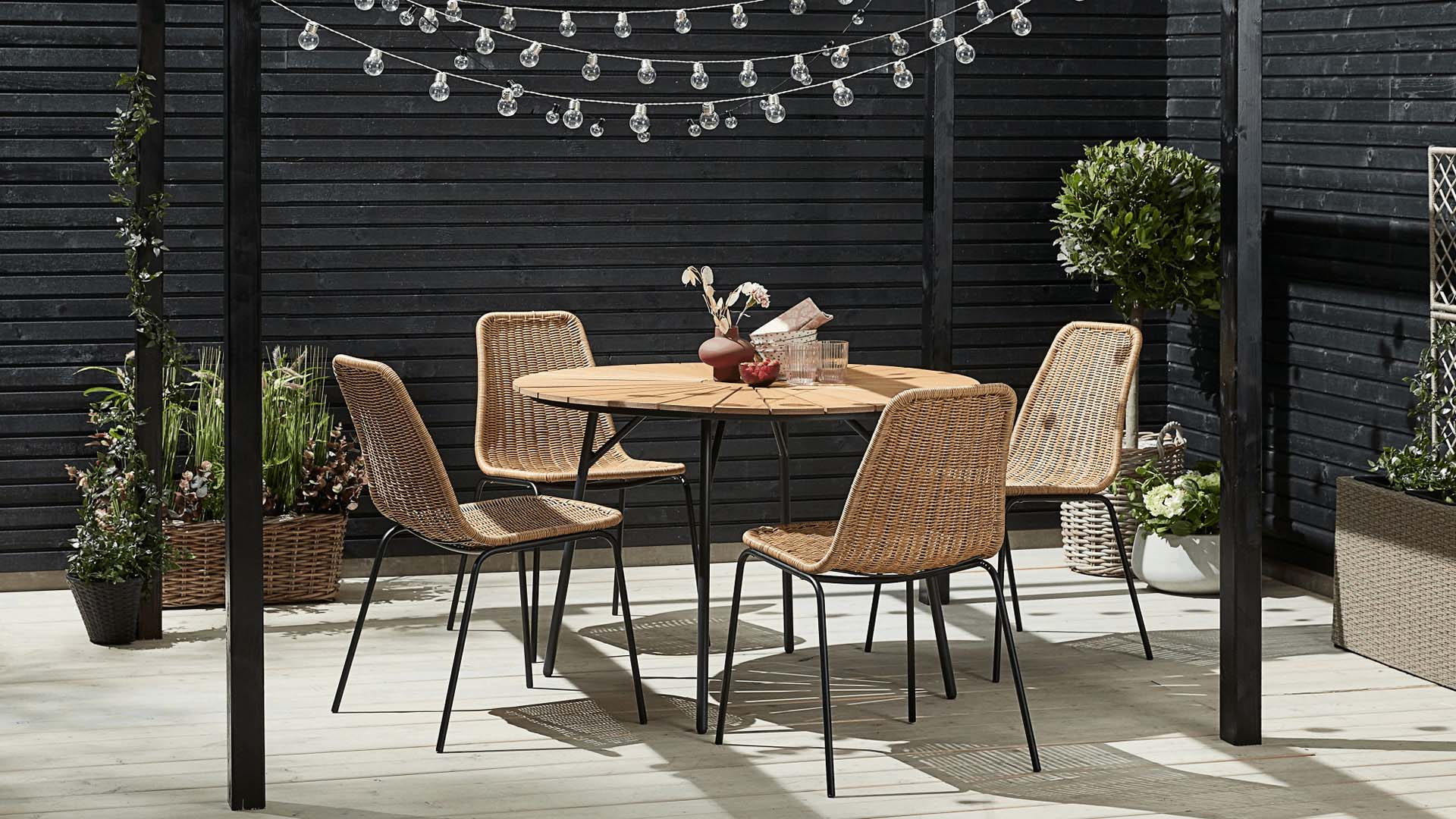 Black metal legs rattan table and chairs on a black pergola surrounded by black side walls on a light wood deck