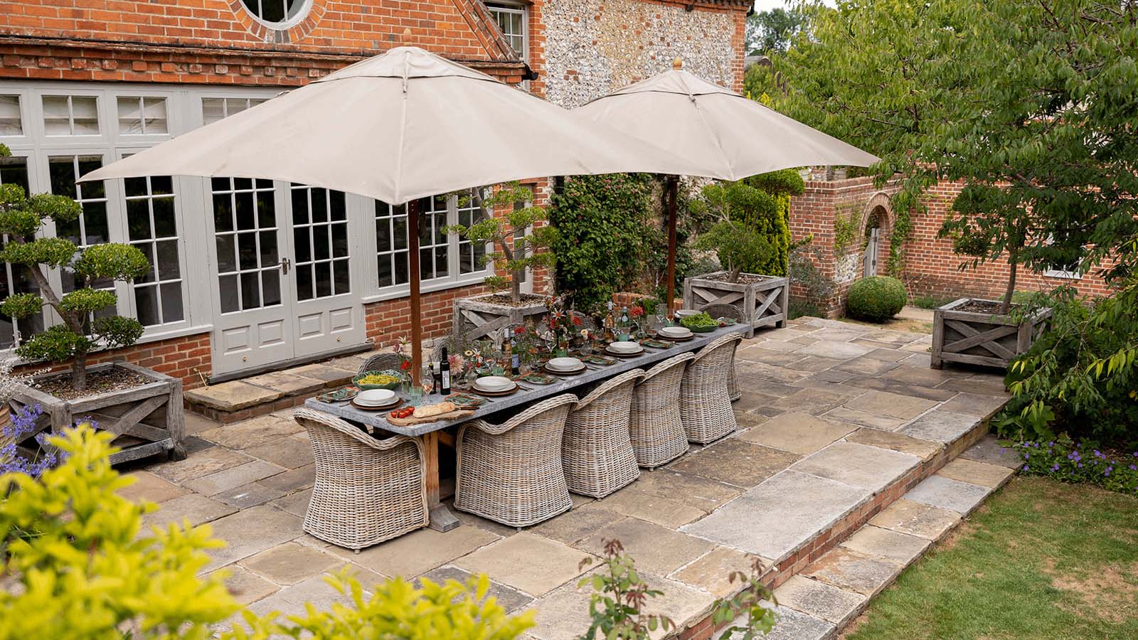 Banquet-style table with comfortable chairs and two parasols in a stone patio. 