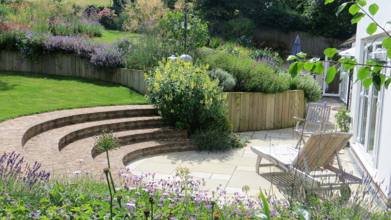 A patio area with curved steps leading up to a lawn, surrounded by planted areas and a wooden sun lounger.