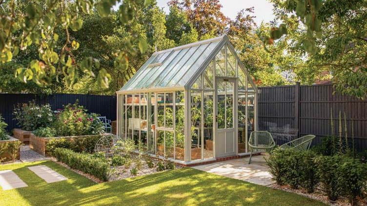 A glass house sits in the corner of a fenced area, surrounded by gravel paths, grass, and plant beds