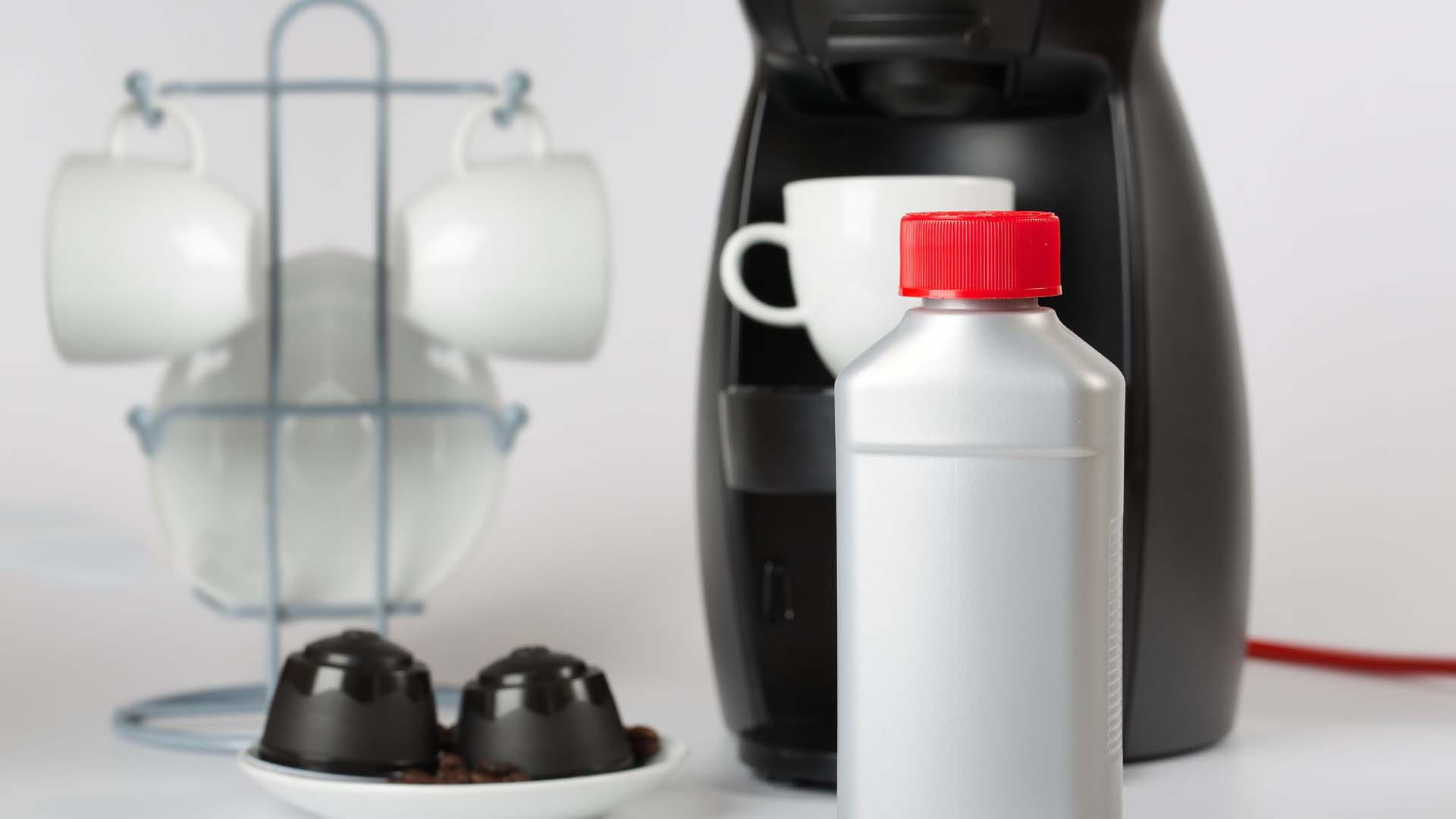 In the foreground is a grey bottle of coffee machine cleaner, surrounded by a black coffee machine, rack of white mugs and plate of coffee pods