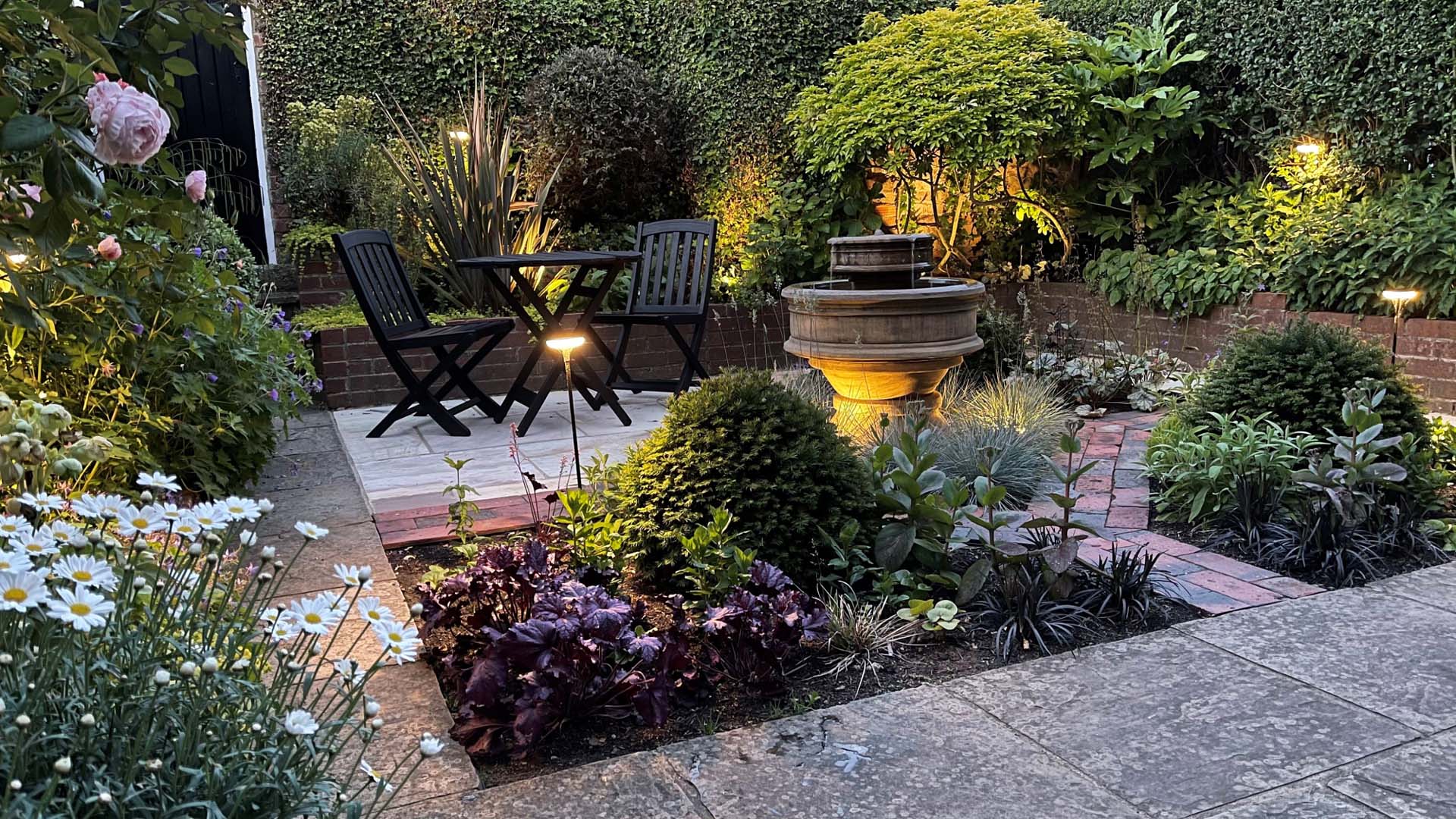 Small stone patio surrounded by evergreen climbers, boundary hedging with a 2 black chairs and small round table