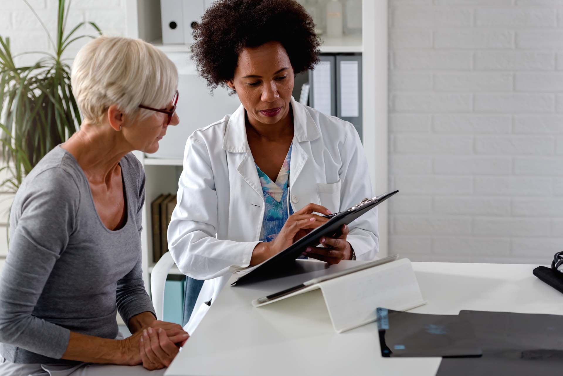 An older woman with cropped blonde hair sits at a desk with a female doctor wearing a white coat, both looking at a clipboard