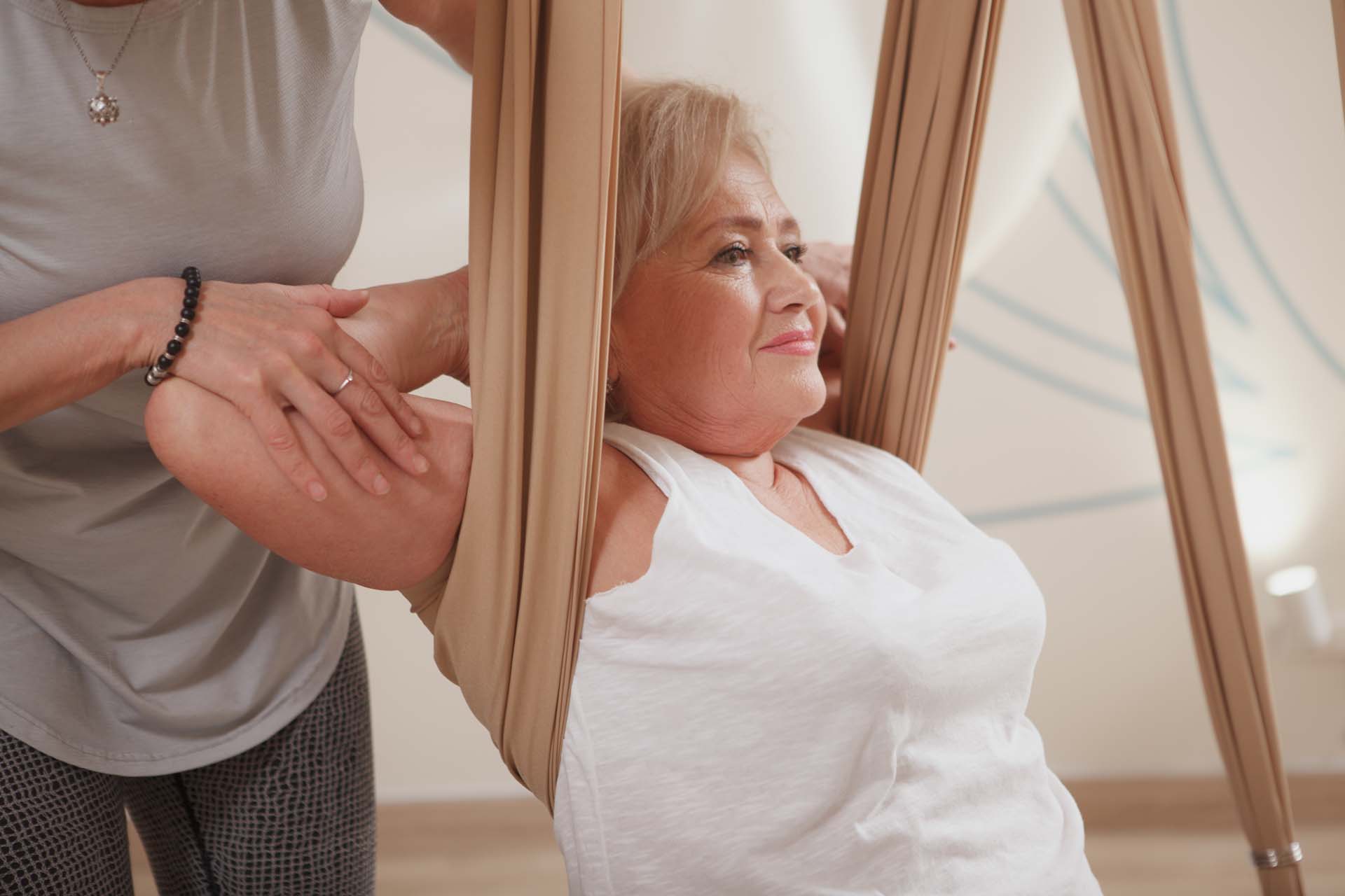 A woman in a white top practicing aerial yoga with silks supporting under her arms, while another woman adjusts her posture.