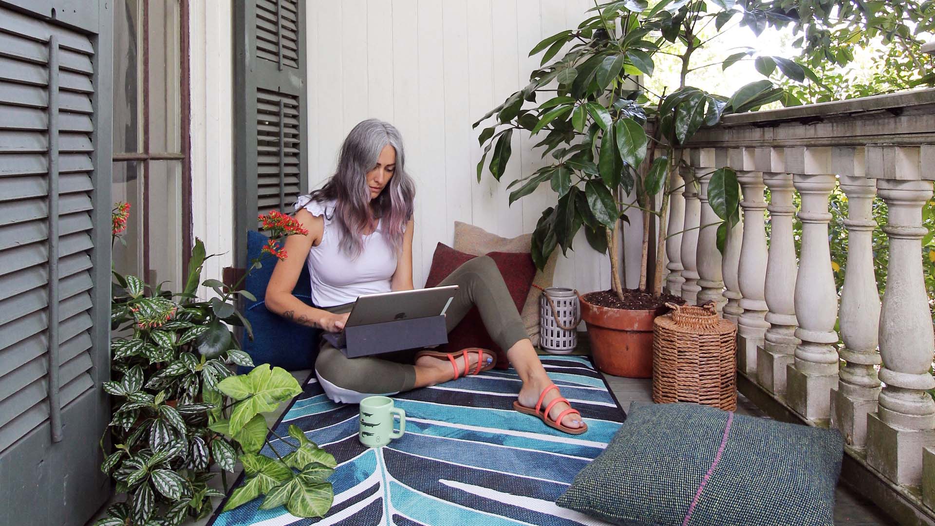 An older lady works on a laptop sitting on a rug on her balcony