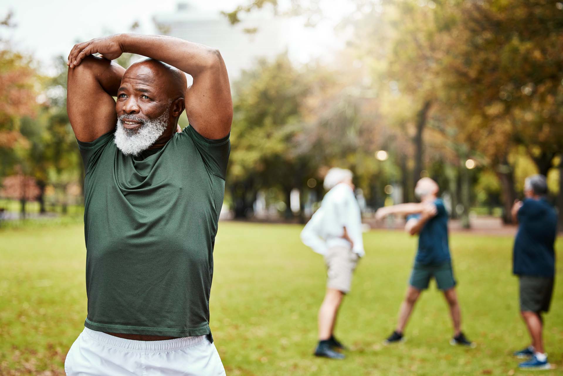A black man with a white beard stretches his arm muscles in a park with other stretching people visible in the background.
