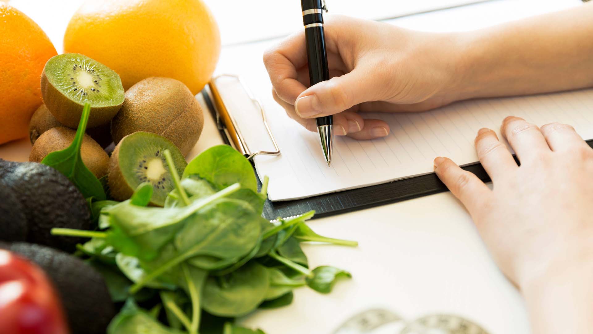 A hand holding a pen writing a shopping list next to some fruit and veg