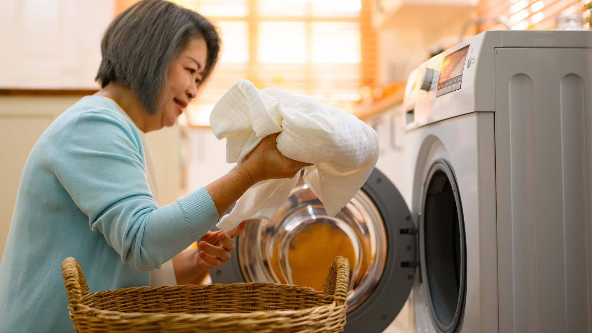An older woman with short dark hair loads a towel into a washing machine from a wicker basket.