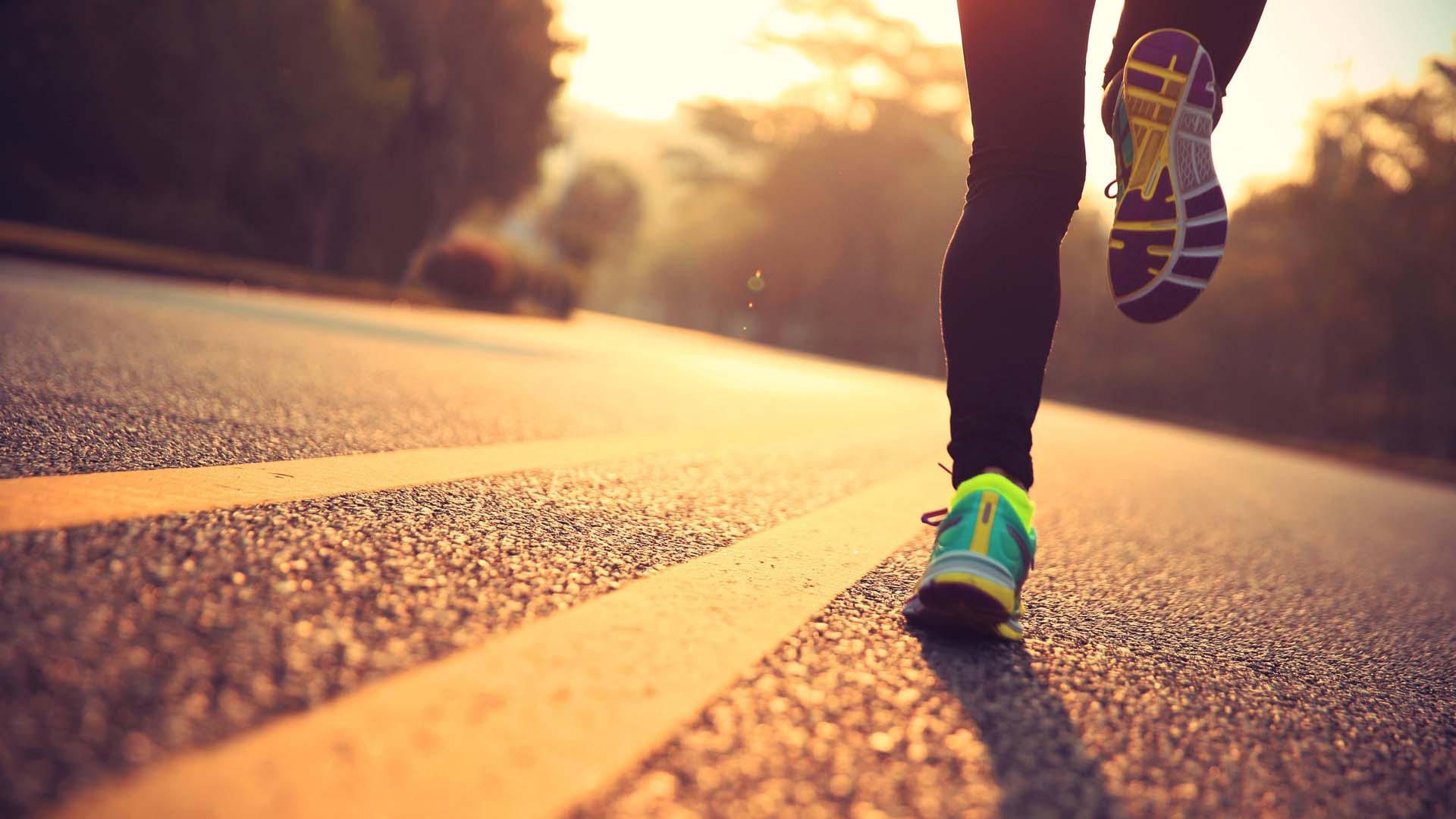 Photo taken behind runner on road with the bottom of their legs showing and the sun rising in front. 