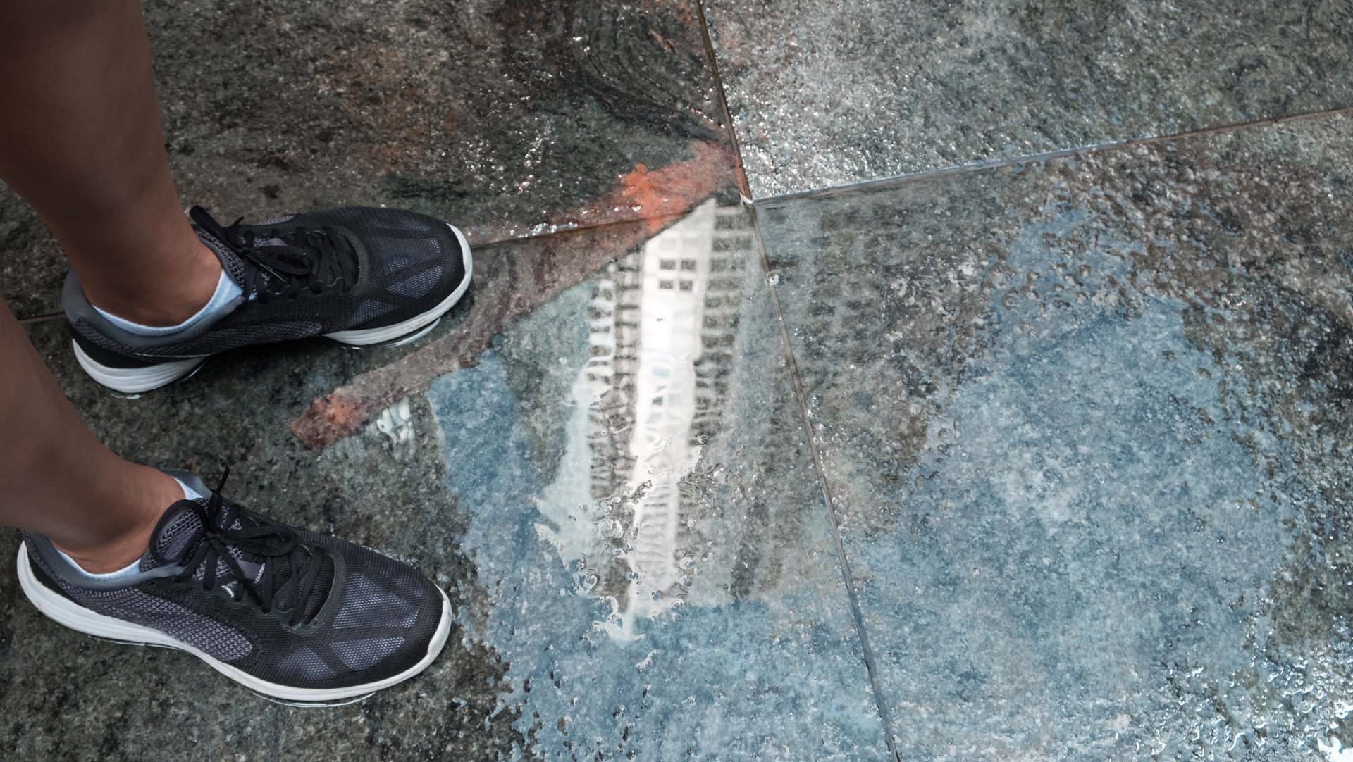 Runner standing on pavement, puddle on floor with high rise building reflected in it