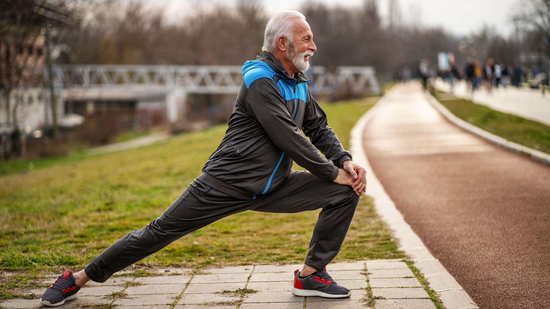 An older man with white hair and facial hair wearing fitness clothing performs a leg stretch outdoors next to a stretch of path