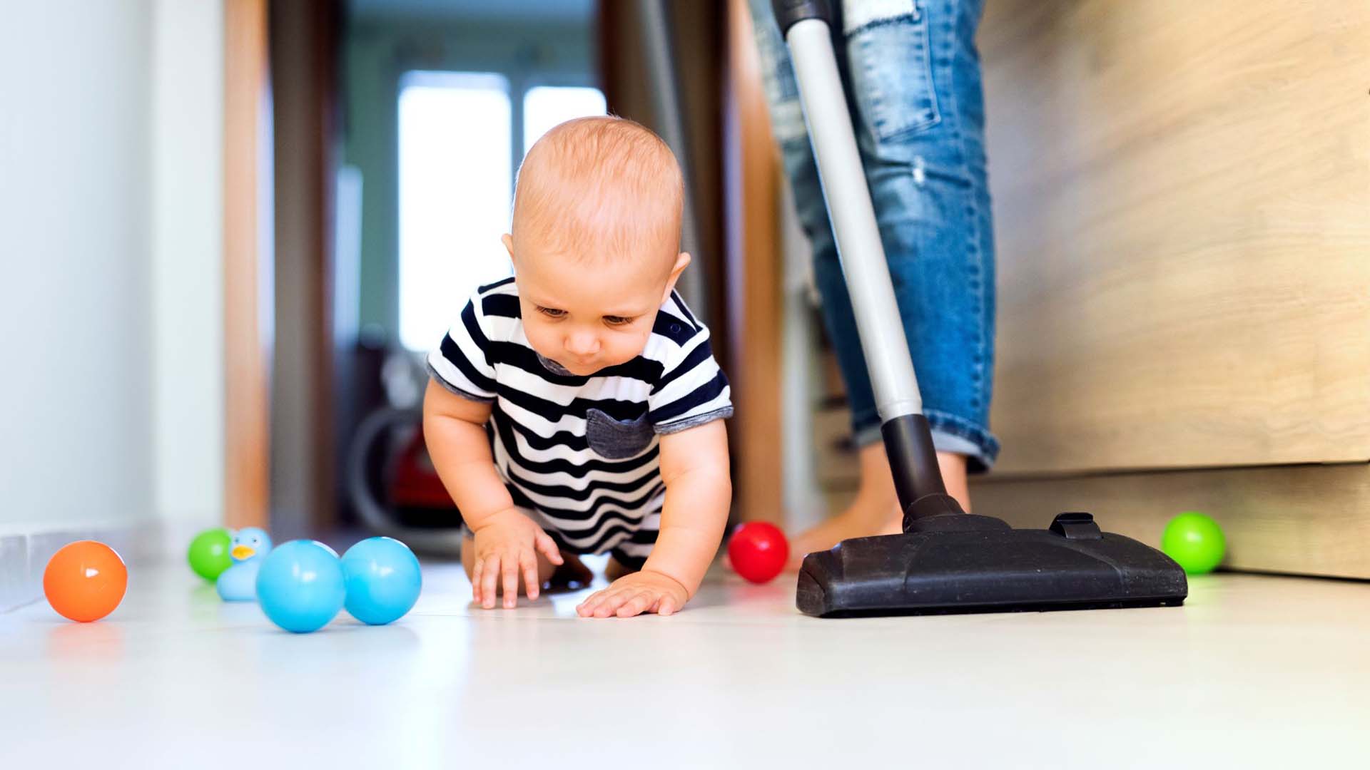 baby sat among toys while an adult vacuums around