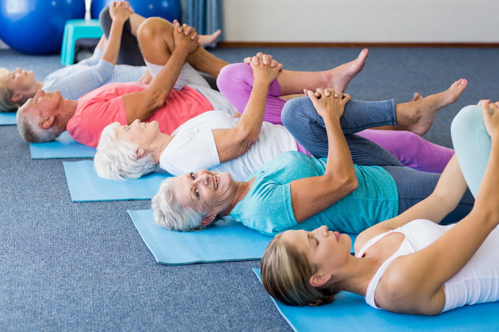A class of middle-aged women and men lying on blue yoga mats in a diagonal line, stretching a knee to their chests. A grey-haired woman smiles at the camera while stretching.