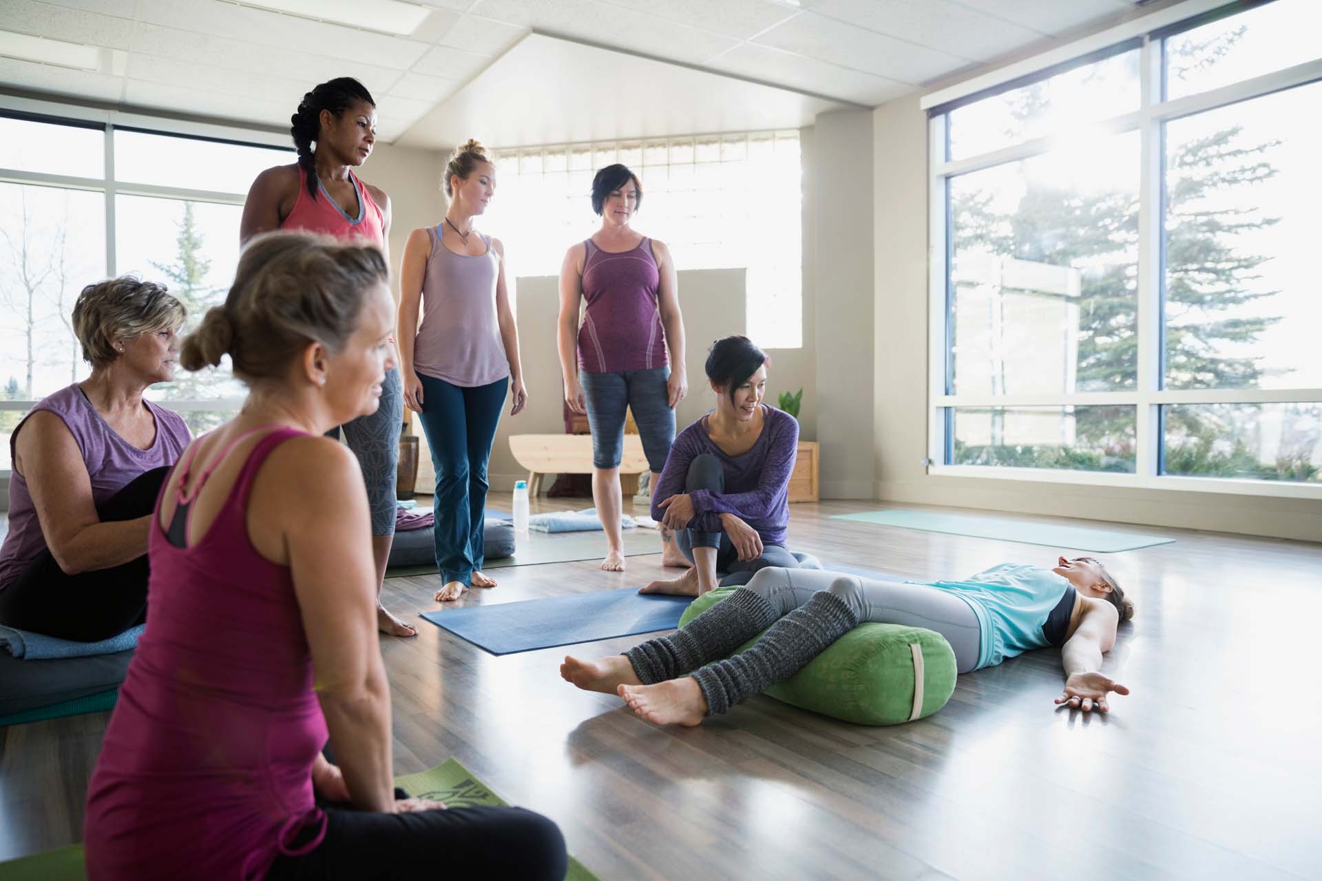 A yoga instructor lying on a yoga mat with a bolster under her knees, demonstrating to a group of women in yoga attire who are standing or sitting in a yoga studio setting.