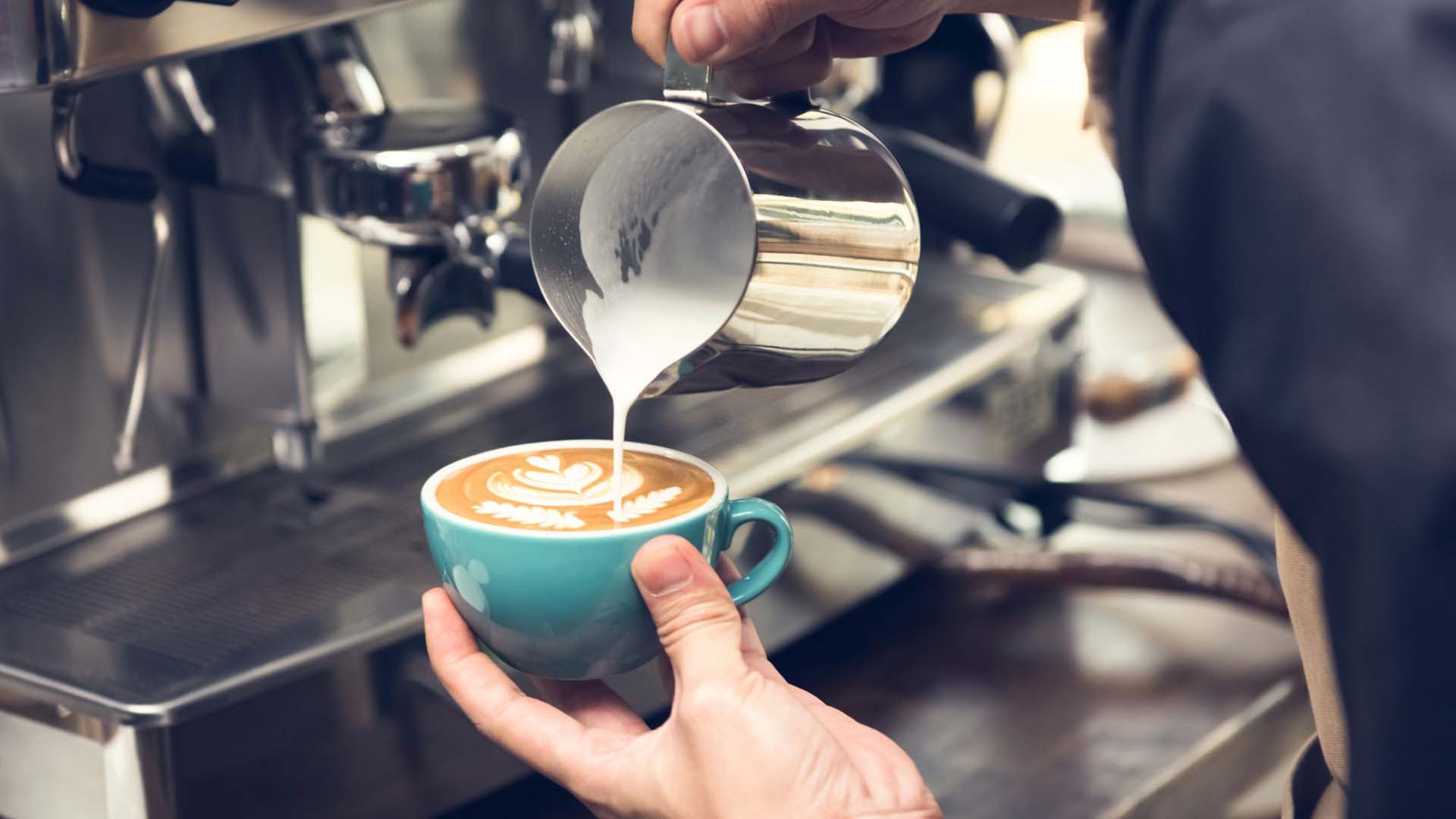 Close-up of a barista pouring frothed milk into a coffee mug in front of a large coffee machine