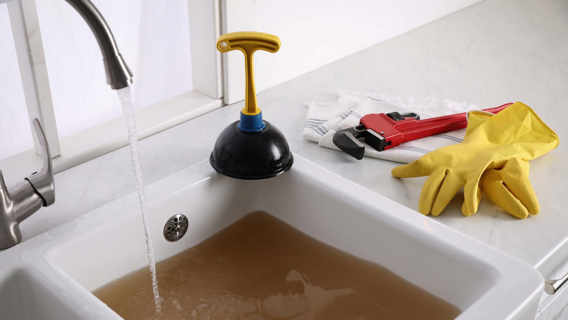 A blocked sink with a plunger and rubber gloves next to it