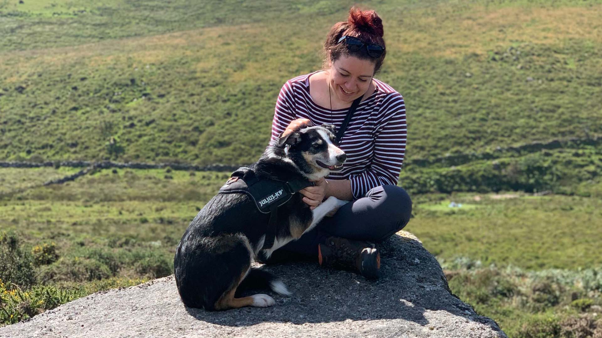 A woman sitting on a large rock with her dog and open countryside views behind them