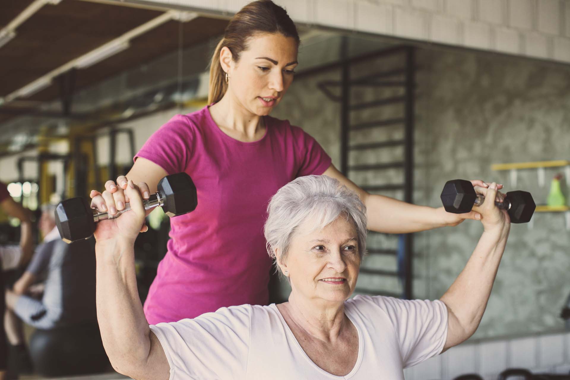 An instructor teaching an older woman to lift weight in a gym